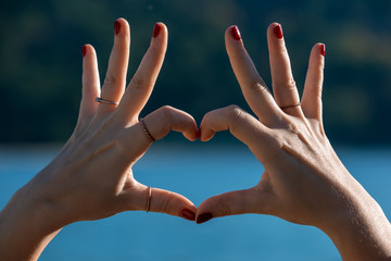 Female hands in heart shape in focus, blue sea and beachfront on background 