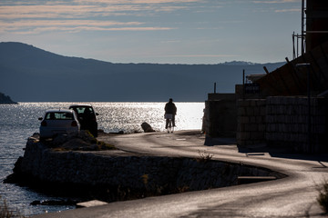 Silhouette of man cycling on beachfront asphalt road, silhouettes of cars on the left side of the road, sunshine glows on sea surface