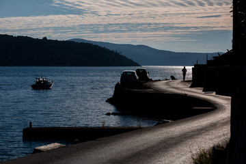 Silhouette of a person walking on beachfront asphalt road, silhouettes of cars on the left side of the road,  partly cloudy sky, contour light
