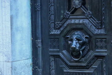 An Egyptian style carved cheetah head door handle with plain ring through its mouth sits on a elaborately decorated Victorian mausoleum metal door in Highgate West Cemetery, London, UK.