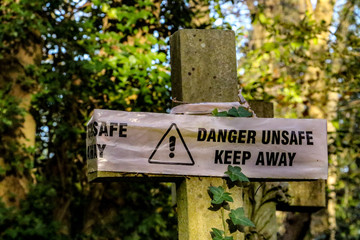 Damaged Victorian stone cross in Highgate Cemetery with danger unsafe keep away sign rapped around it. The Cemetery is one of London's magnificent seven graveyards and is now a unique urban wild area