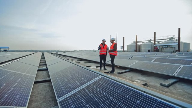 Engineers talk on the solar farm background. Rows of solar panels and two workers in orange uniform and hard hat. Installation of innovative sunny batteries.
