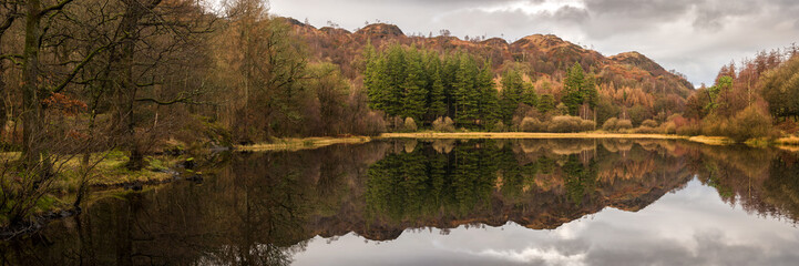 Fototapeta premium Piękny panoramiczny krajobraz wiejski lasów i gór odzwierciedlony w jeziorze w burzliwy dzień na pojezierzu w Anglii.