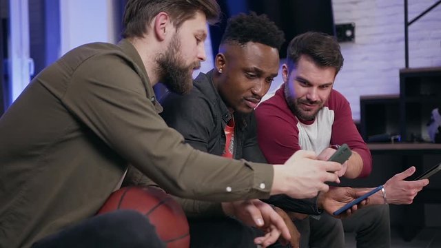 Side view of good-looking serious modern young diverse guys which sitting on sofa and using their phones while watching tv programm
