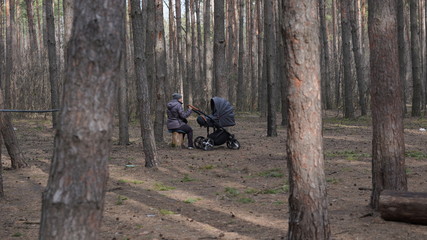 Grandmother on walk with her grandson or granddaughter in forest. Elderly woman with stroller resting on stump in woodland.