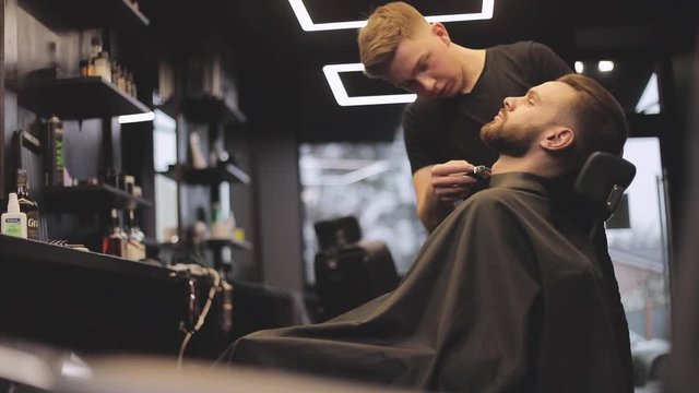 Barber shop concept. Handsome young confident bearded man is sitting on hairstyling chair in the salon while hairdresser makes cool beard and hairstyle for his client.