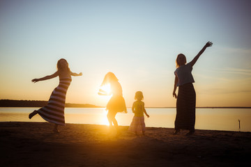 Silhouettes of children and their mothers jumping and having fun on the beach in sunset light. Good mood and pastime among the younger and older generation.