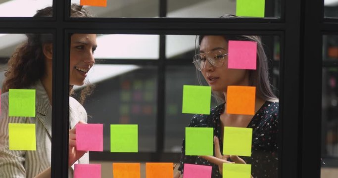 Positive multiethnic young businesswomen managing working process together at glass kanban board, using colorful paper stickers. Smart female diverse colleagues discussing project tasks in office.