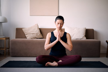 Young attractive woman practicing yoga, sitting in Padmasana, exercise, Lotus pose, namaste, working out, wearing sportswear, black t-shirt, pants, indoor full length