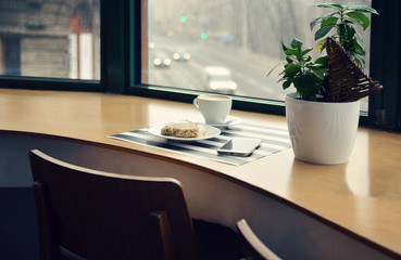 Wooden office table with big window with phone, plant, cup of coffee and cookie. Copy space. Modern interior of workplace in cafe.