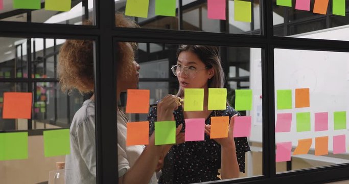 Focused young vietnamese project manager discussing working processes with motivated african american teammate near window kanban board. Multiracial colleagues writing creative ideas on sticky papers.