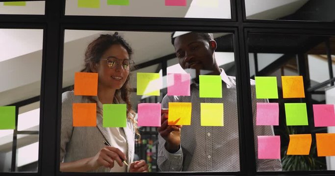 Happy young male african american company employee managing project processes with caucasian female colleague in eyeglasses at kanban glass board, using colorful sticky papers in modern office.