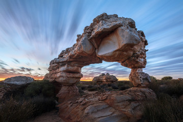 A dramatic landscape photograph of an incredible rock arch before sunrise, with fast moving clouds against a blue sky, taken in the Cederberg mountains, South Africa.