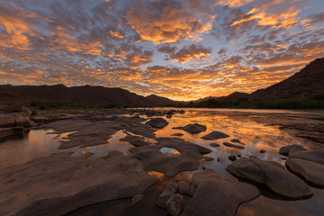 Fototapeta na wymiar A beautiful landscape of a golden sunset over the mountains and calm waters of the Orange River, with dramatic yellow clouds reflecting in the water’s surface, taken in the Richtersveld South Africa.