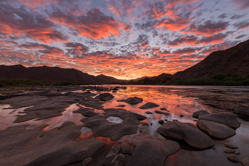 A beautiful landscape of a golden sunset over the mountains and calm waters of the Orange River, with dramatic orange clouds in the sky, taken in the Richtersveld National Park, South Africa.