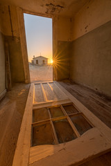 A dramatic vertical photograph inside an abandoned house at sunset, with an old door lying on the floor and a sunburst and house outside, taken in the ghost town of Kolmanskop, Namibia.