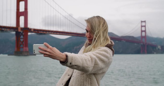 A young woman using her phone taking a video with the famous Golden Gate Bridge in the background.
