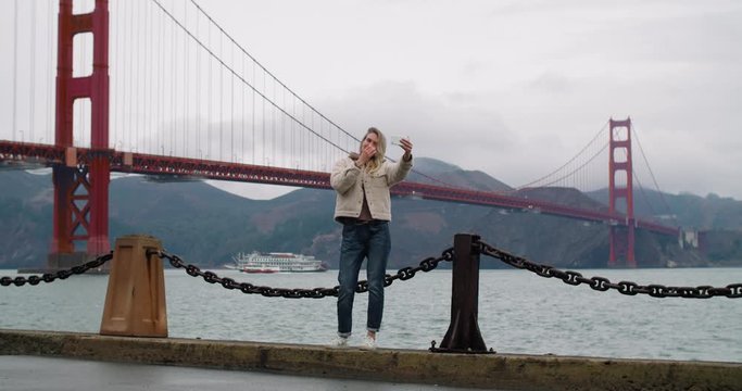 A young woman taking a selfie photo using her phone with the famous Golden Gate Bridge in the background.