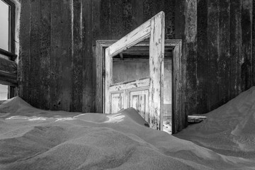 A black and white photograph inside an abandoned house with an open door submerged in the rippled desert sand, taken in the ghost town of Kolmanskop, Namibia.