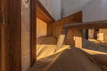 A photograph inside an abandoned house with an open door submerged in the rippled desert sand and golden sunlight streaming in, taken in the ghost town of Kolmanskop, Namibia.