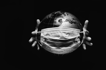 Hands of little child holding globe sphere, planet map covered with medical protective mask isolated on black background. Concept of COVID-19 pandemic infection. Black and white image