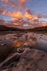 Fototapeta na wymiar A vertical landscape of a golden sunset over the mountains and calm waters of the Orange River, with dramatic orange clouds reflecting in the water’s surface, taken in the Richtersveld South Africa.