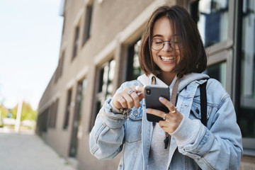 Youth, technology and spring concept. Cheerful pretty young woman in glasses, denim jacket,...