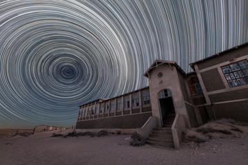 Fototapeta premium A spooky and dramatic night sky photograph of the old hospital building with circular star trails and desert sand, taken in the ghost town of Kolmanskop, Namibia.