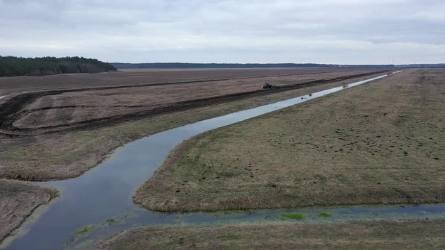 Two parts of field-before and after plowing, and working tractor in background. Irrigation canal near the field. An open channel for watering fields. Cloudy day. Agricultural landscape from the air.