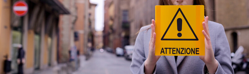 Coronavirus. Girl holds a yellow sign in her hands with the text Atenzione in Italian. The concept of pandemic and infectious diseases worldwide. Girl on the street of Italy in the city. Banner long