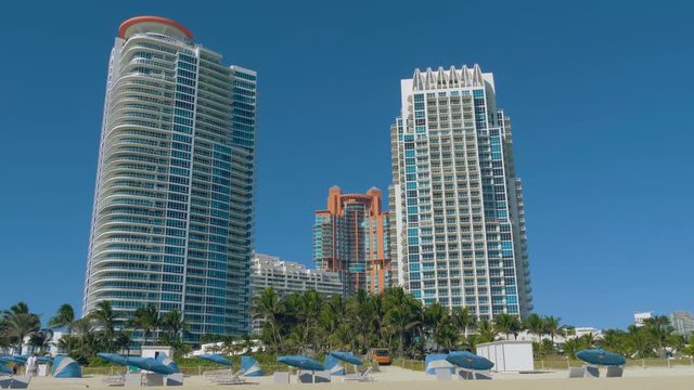 Miami Beach in Florida with luxury apartments and beach umbrellas. Residential buildings, skyscrapers on the shore of Miami Beach on a sunny day. Park area with palm trees near buildings.