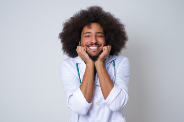 African American doctor man keeps hands pressed together under chin, looks with happy expression, has toothy smile, isolated over gray background