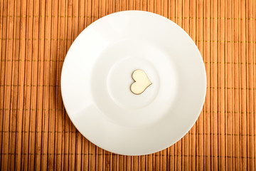 Top view of one white bowl with a small wooden heart on a table, in warm light, brown monochrome indoor background photographed with selective focus