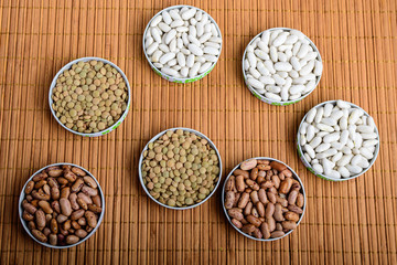 Top view of seven bowls with organic fresh red and white beans and lentils on a wooden table displayed as a flower in warm light, brown monochrome indoor background photographed with selective focus