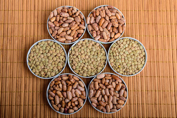 Top view of seven bowls with organic fresh red beans and lentils on a wooden table displayed as a flower in warm light, brown monochrome indoor background photographed with selective focus