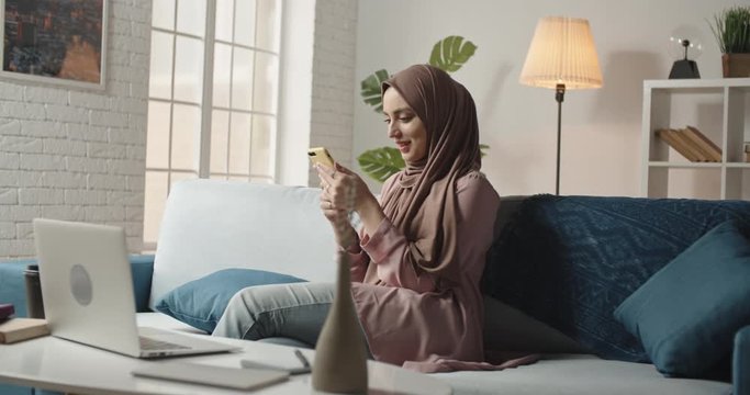 Beautiful young muslim girl in traditional hijab scarf is using her smartphone while working on project and positively smiling. Successful female freelancer or university student studying 4k footage