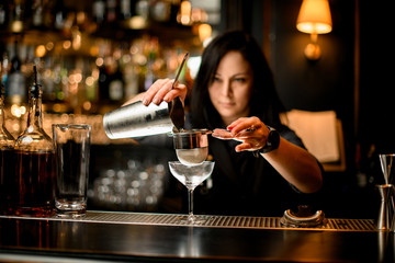 brunette woman barman gently pours cocktail from shaker into glass using sieve.