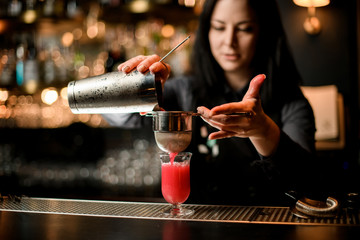 Fototapeta na wymiar woman bartender professionally pours cocktail from shaker into glass using sieve.