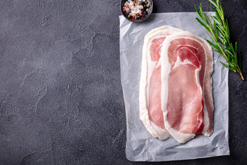 Raw bacon with rosemary and spices slices on parchment paper. Grey slate background. Copy space....