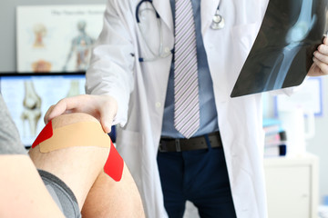Close-up of smart physician in white uniform and striped tie checking damaged ankle. Troubled joint bound with protective red patches to prevent blood infection. Medical treatment concept