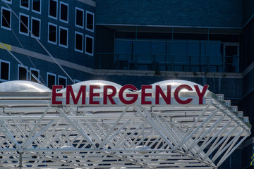 Obraz premium An emergency sign outside a hospital. The illuminated red letters spell emergency and is affixed to white metal beams. There are windows in the background of a large hospital building. 