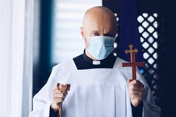 Man priest in medical mask praying with cross and rosary.