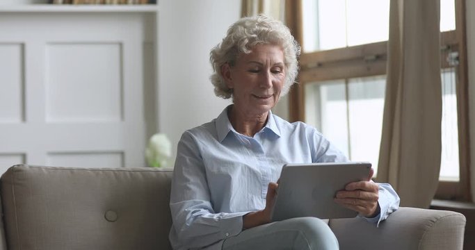 Smiling 60s mature older woman sitting on comfortable couch, using digital tablet. Middle aged granny using computer applications, chatting in social networks with friends, shopping online at home.