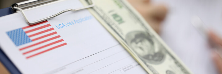 Close-up view of persons hand holding clipboard with usa visa application form and cash money to...