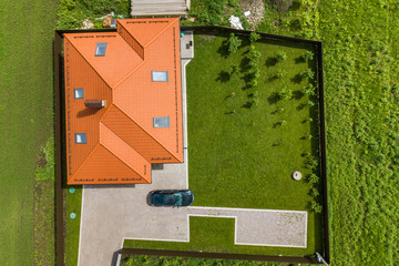 Aerial top view of house shingle roof with attic windows and black car on paved yard with green grass lawn.