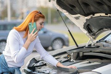 Young woman standing near broken car with popped hood talking on her mobile phone while waiting for help.