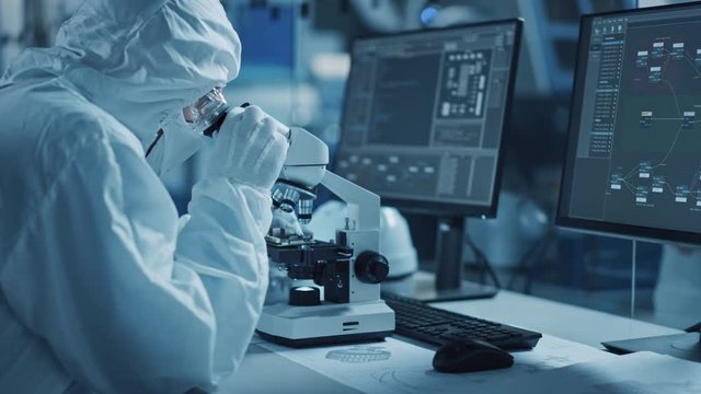 Research Factory Cleanroom: Engineer / Scientist wearing Coverall, Gloves Uses Microscope to Inspect Samples, Developing High Tech Modern Technology for Medical and High Precision Electronics Industry