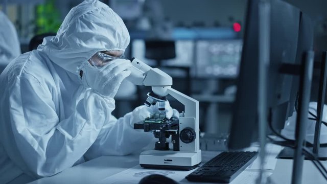 Research Factory Cleanroom: Engineer / Scientist wearing Coverall, Gloves Uses Microscope to Inspect Samples, Developing High Tech Modern Technology for Medical and High Precision Electronics Industry