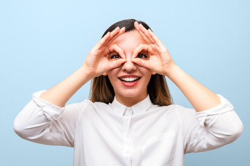 Fototapeta premium vision aids. young woman holding her hands over her eyes imitating glasses and smiling cheerfully against blue background