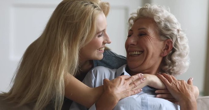 Attractive young woman cuddling smiling elderly mother, posing for photo together. Head shot close up happy mature senior mother feeling joyful with grown up daughter sitting on comfortable couch.